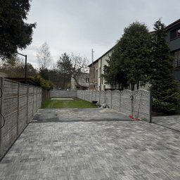 Oleksandr Chyplyk - Szara kostka brukowa na podjeździe, otoczona betonowym ogrodzeniem i zielonym trawnikiem. Widoczne narzędzia brukarskie przy ogrodzeniu. Pochmurne niebo w tle.