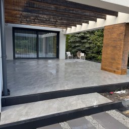 RekBruk - Nowoczesny taras z szarych płyt, drewniana pergola i elementy dekoracyjne. Widok na ogród z krzesłami. Minimalistyczny design, świeżo po deszczu.