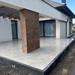 RekBruk - Nowoczesny taras z szarych płyt, obrzeża z czarnej kostki. Drewnopodobny filar i jasna elewacja domu w tle. Minimalistyczny design.