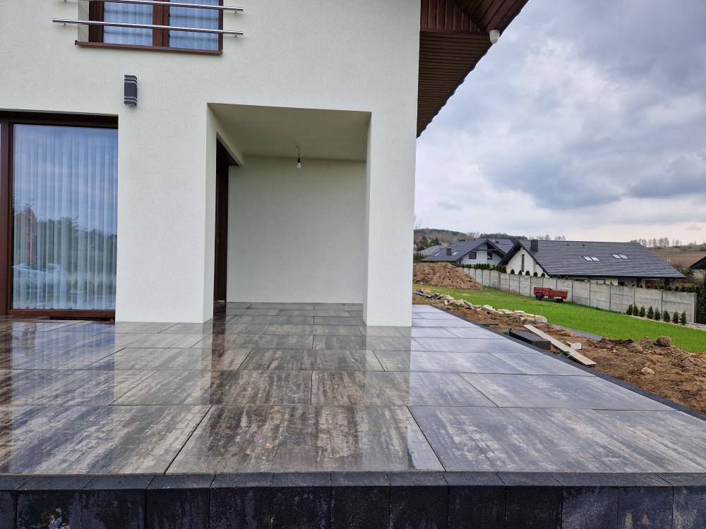 Nowoczesny taras z szarej kostki brukowej o nieregularnym wzorze, przylegający do białego budynku z dużymi oknami i widokiem na ogród w pochmurny dzień.