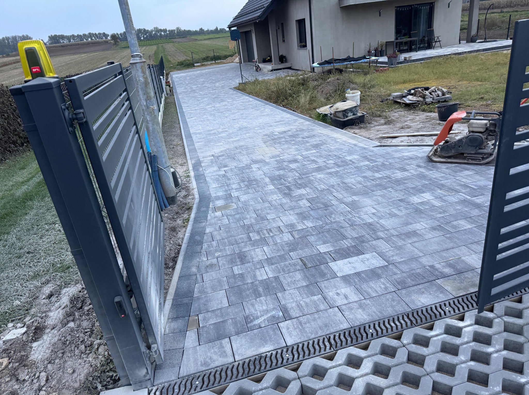 Nowoczesny podjazd z szarej kostki brukowej, widok zza otwartej, metalowej bramy wjazdowej. W tle dom jednorodzinny i zielone pola.