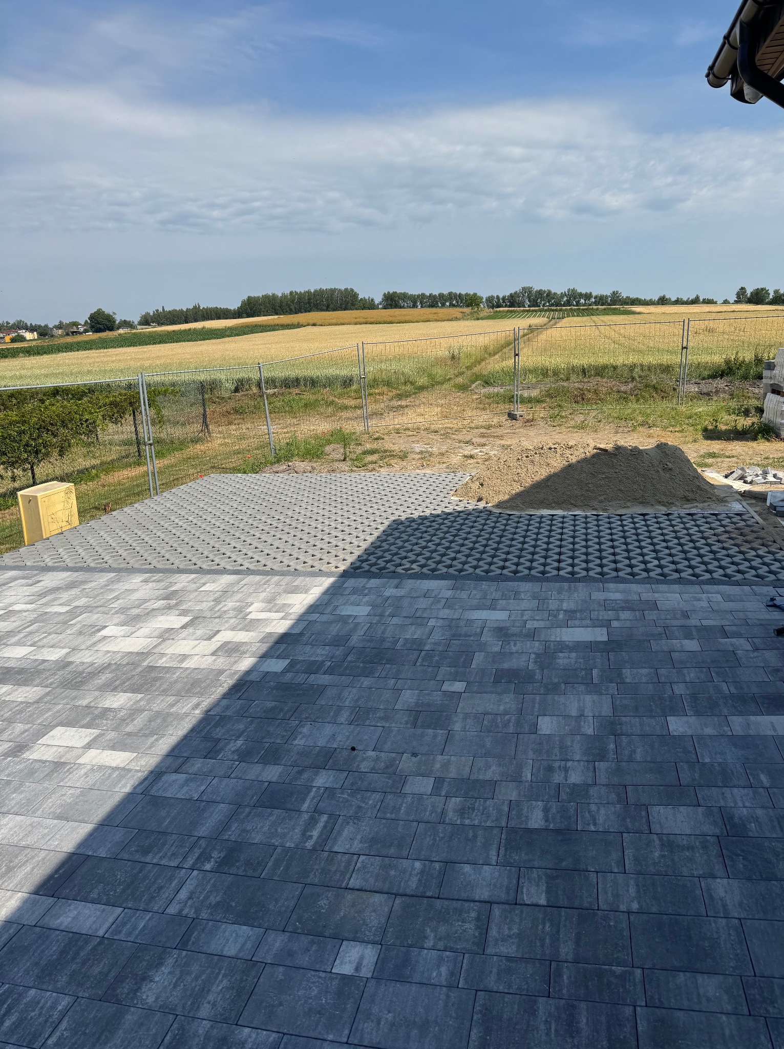 Fragment układanej nawierzchni z kostki brukowej w trzech wzorach, z widokiem na pole i błękitne niebo w tle. Częściowo ułożona kostka, piasek i paleta z kostką.