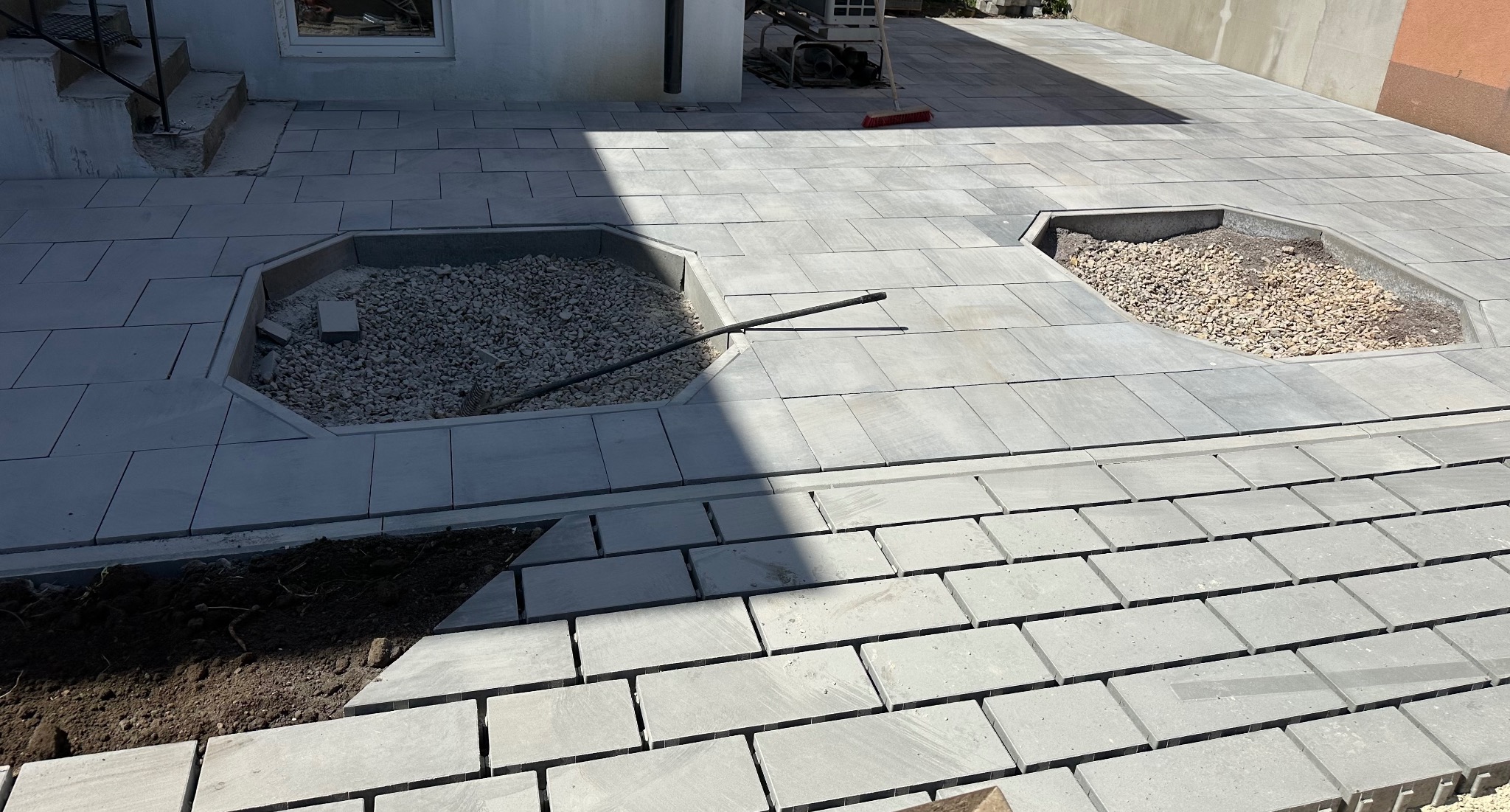 Szara kostka brukowa ułożona częściowo na posesji, z dwoma miejscami na rośliny wypełnionymi żwirem. Widoczne narzędzie i fragment budynku w tle.