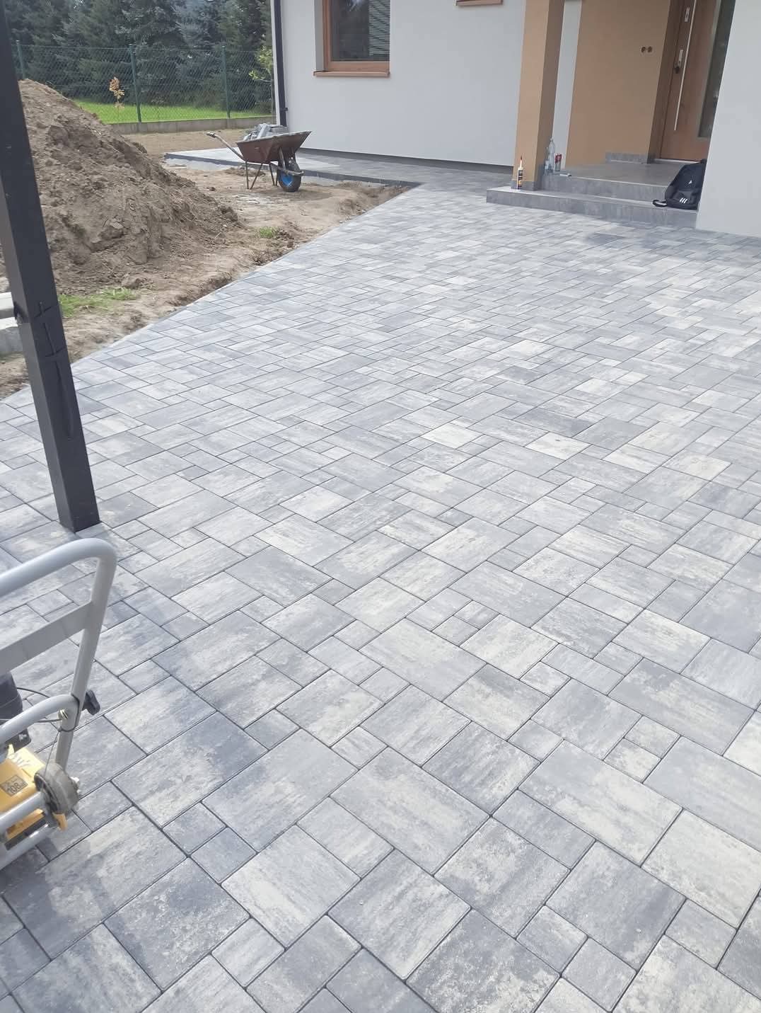 Ułożona szara kostka brukowa na podjeździe domu jednorodzinnego. Widoczna ubijarka, hałda ziemi i taczka. Nowoczesny wzór kostki.