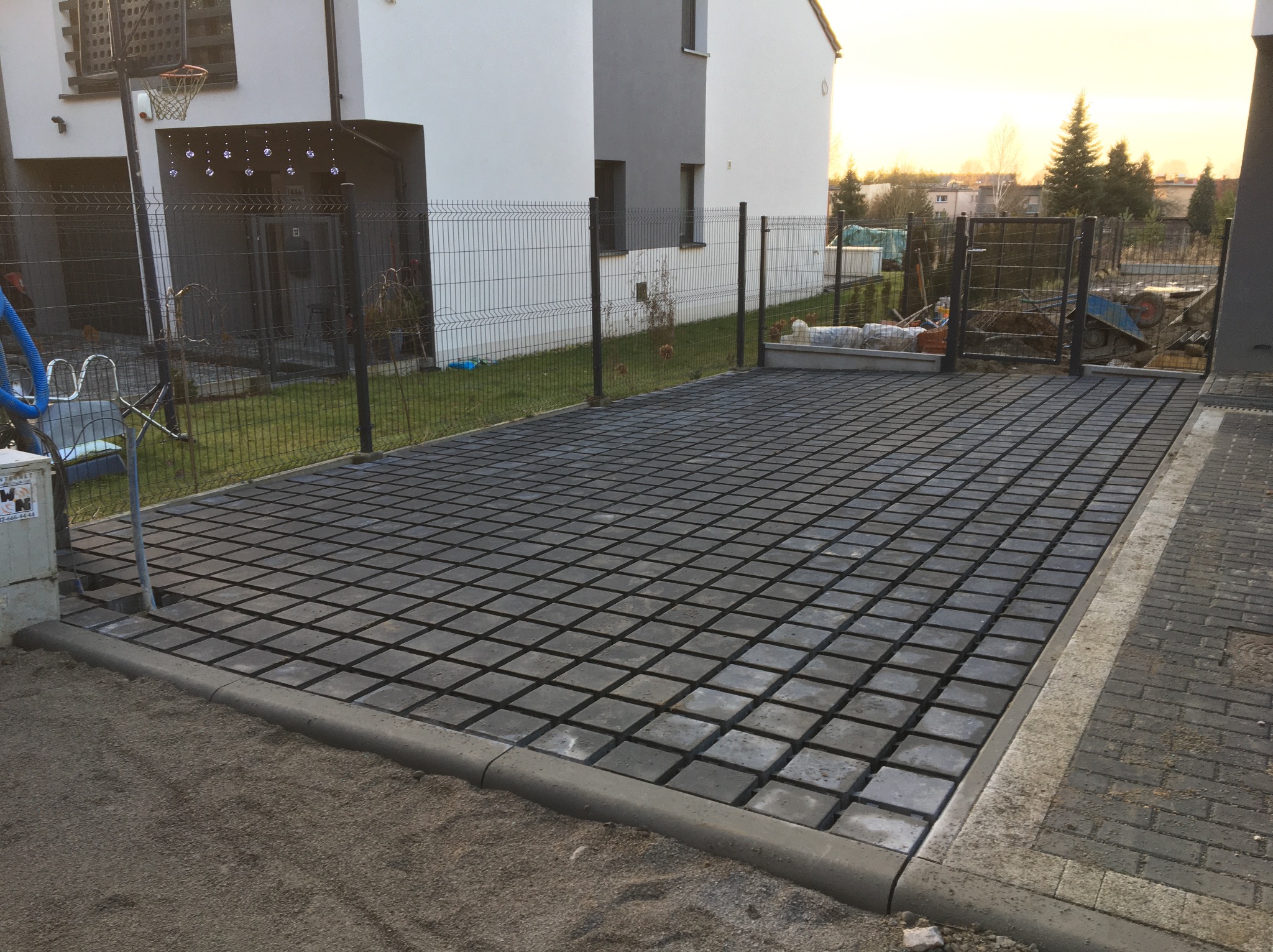 Ułożona, szara kostka brukowa na podjeździe domu jednorodzinnego, oddzielona obrzeżem. W tle widoczny ogród i częściowo sprzęt budowlany.