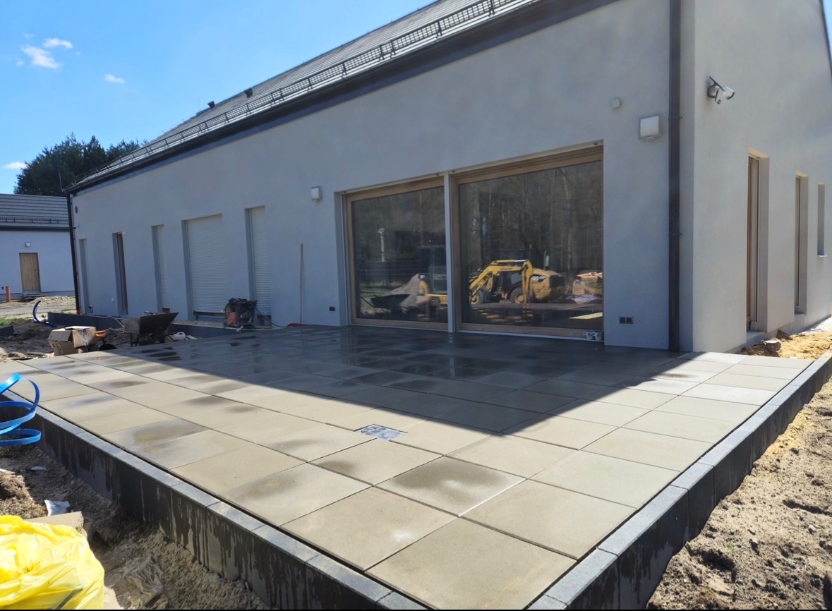 Nowoczesny taras z jasnej kostki brukowej, obrzeża z ciemnej kostki. W tle budynek z dużymi oknami, w których odbija się krajobraz i sprzęt budowlany.