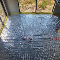 INSTALATORSTWO I MONTAŻ Zbigniew Łudzeń - Rozłożona instalacja ogrzewania podłogowego na folii izolacyjnej, widoczne rury grzewcze w kolorze białym i czerwonym, zabezpieczone taśmą żółtą przy ścianie, w tle widok przez okno na plac budowy.