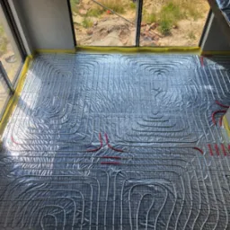 Rozłożona instalacja ogrzewania podłogowego na folii izolacyjnej, widoczne rury grzewcze w kolorze białym i czerwonym, zabezpieczone taśmą żółtą przy ścianie, w tle widok przez okno na plac budowy.