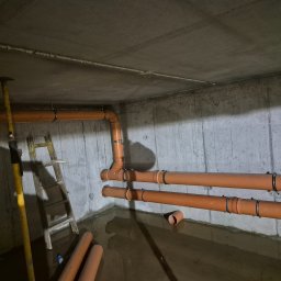 INSTALATORSTWO I MONTAŻ Zbigniew Łudzeń - Pomarańczowe rury kanalizacyjne zamocowane do betonowej ściany w surowym pomieszczeniu, częściowo zalane wodą, widoczna drabina i podpora stropowa.