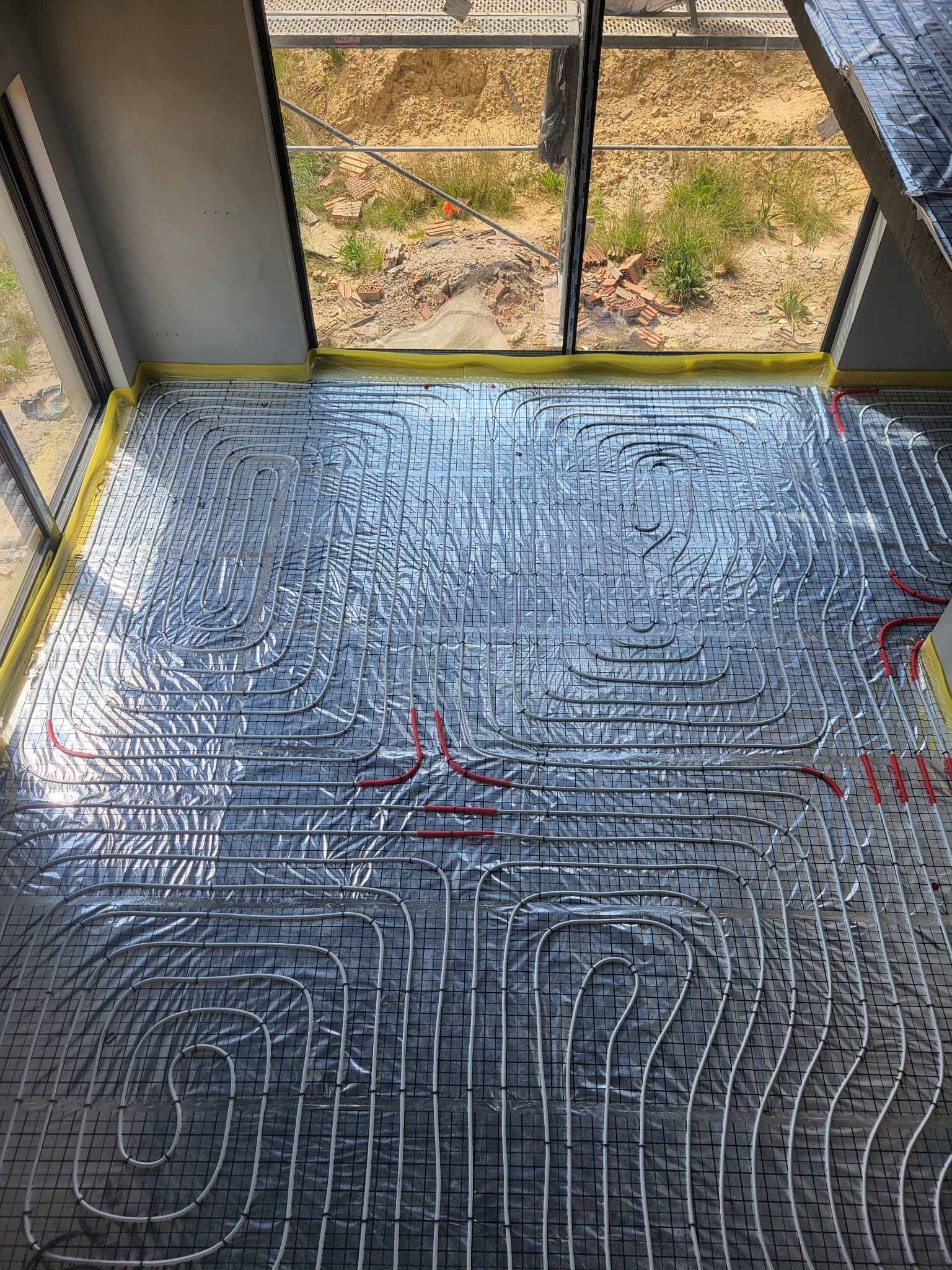 Rozłożona instalacja ogrzewania podłogowego na folii izolacyjnej, widoczne rury grzewcze w kolorze białym i czerwonym, zabezpieczone taśmą żółtą przy ścianie, w tle widok przez okno na plac budowy.