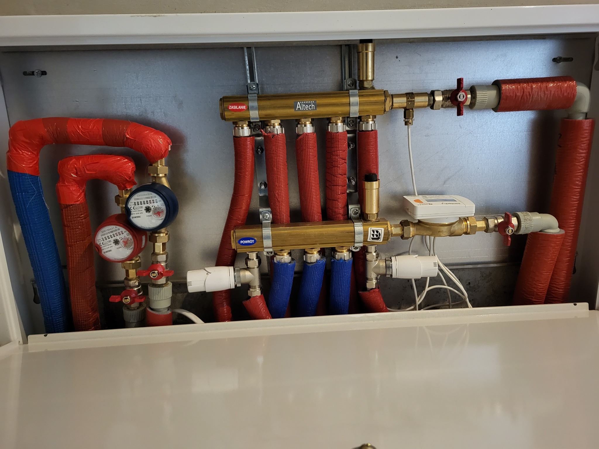 Rozdzielacz instalacji grzewczej w szafce, z widocznymi rurami w izolacji czerwonej i niebieskiej, oraz licznikiem zużycia, zamontowany na metalowej płycie wewnątrz szafki.