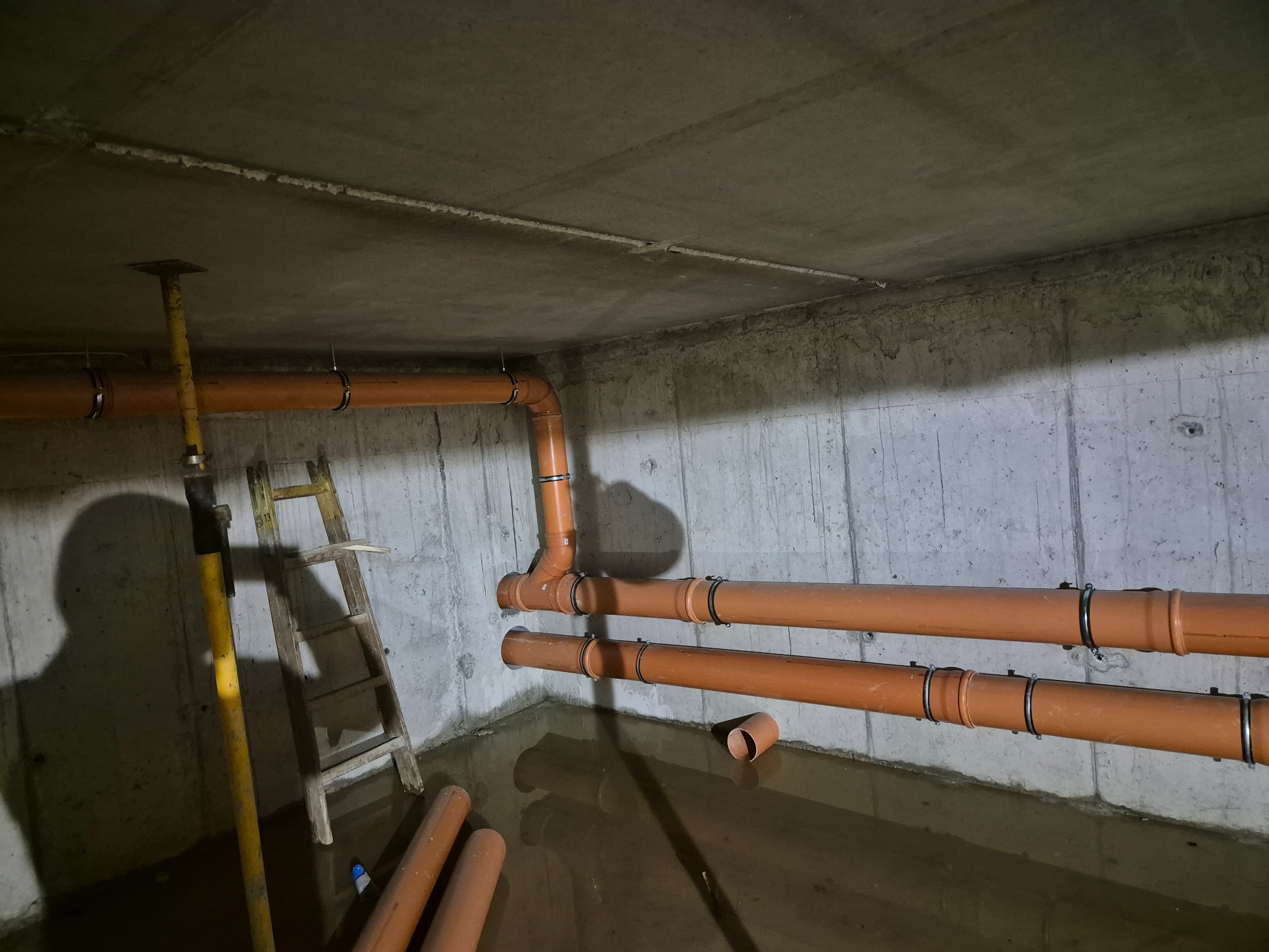Pomarańczowe rury kanalizacyjne zamocowane do betonowej ściany w surowym pomieszczeniu, częściowo zalane wodą, widoczna drabina i podpora stropowa.