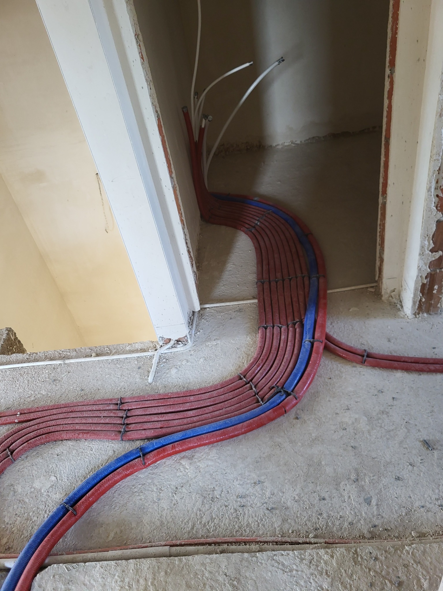 Rury grzewcze podłogowe ułożone w pętlę, z widocznymi rurami czerwonymi i niebieskimi, przymocowane czarnymi opaskami, na surowym podłożu w trakcie instalacji.
