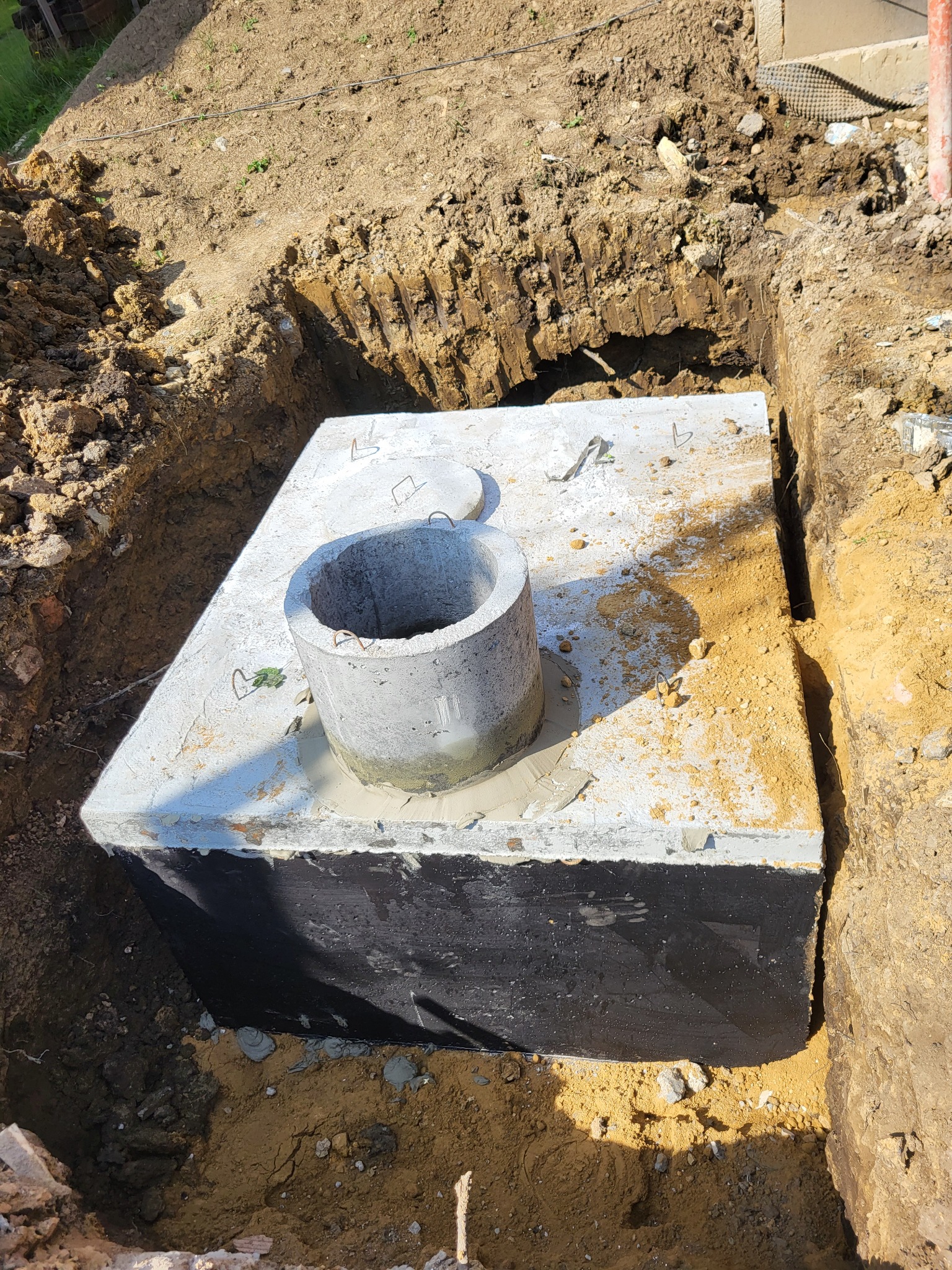 Betonowy zbiornik, prawdopodobnie szambo lub studzienka, umieszczony w wykopie ziemnym, zabezpieczony czarną folią hydroizolacyjną.