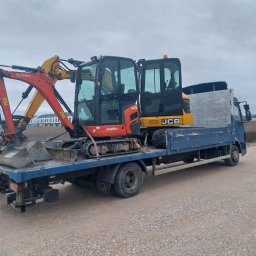 MI-TRANS Mirosław Mitrowski - Niebieska ciężarówka z platformą przewozi pomarańczowo-żółtą minikoparkę Kubota JCB na szarej, żwirowej drodze; widoczne budynki w tle.