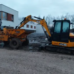 Żółta minikoparka JCB i wywrotka Thwaites w błotnistym terenie budowy, na tle nowoczesnego budynku z płaskim dachem i drzew bez liści.