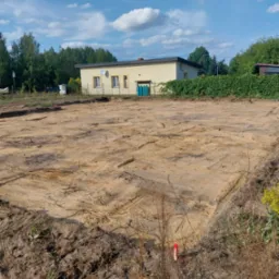 Przygotowany, wyrównany teren pod budowę, widoczne ślady po maszynach, w tle dom jednorodzinny i zieleń.