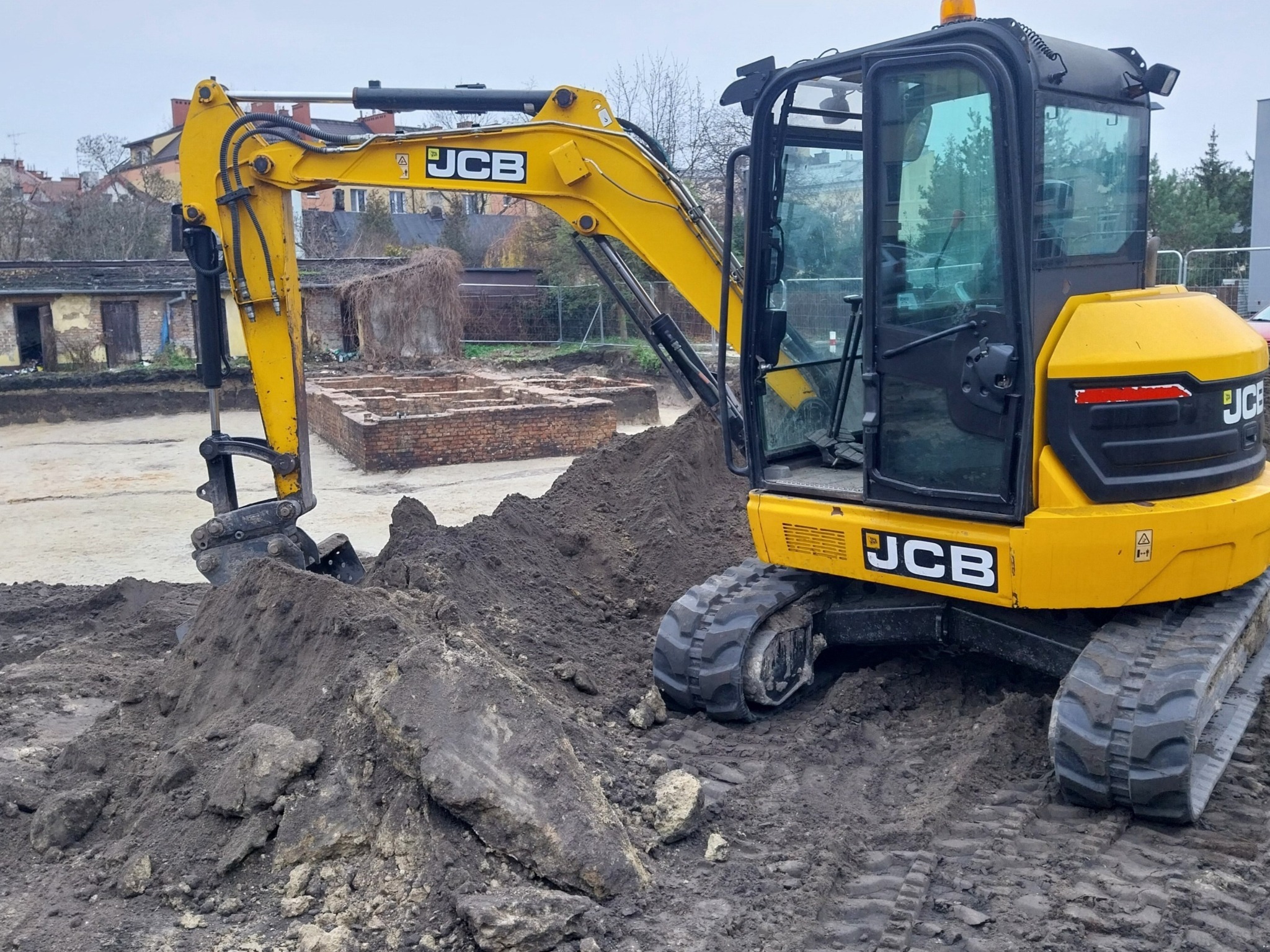 Żółta minikoparka JCB nabiera ziemię z wykopu, w tle widoczne fundamenty budynku.