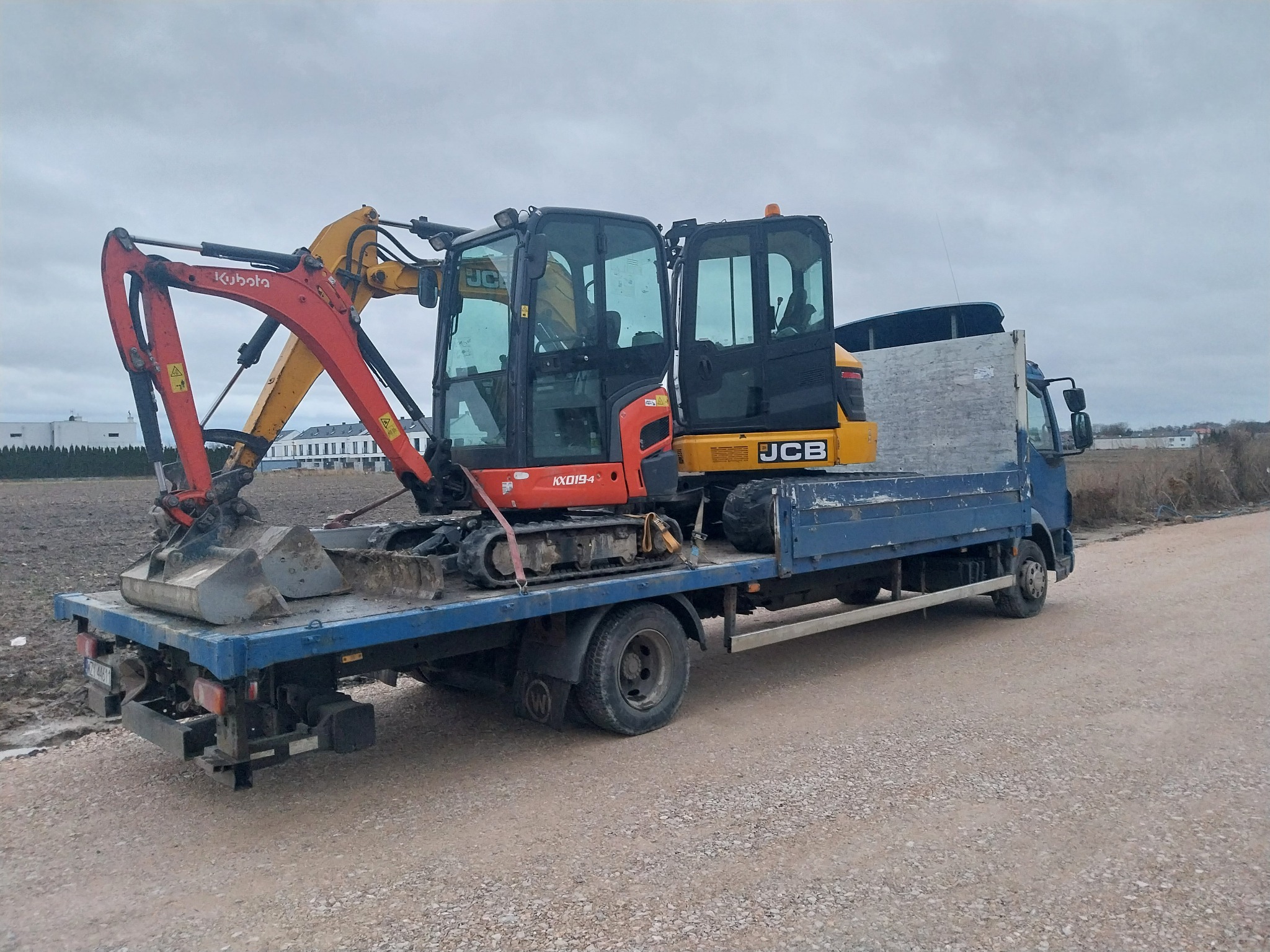 Niebieska ciężarówka z platformą przewozi pomarańczowo-żółtą minikoparkę Kubota JCB na szarej, żwirowej drodze; widoczne budynki w tle.