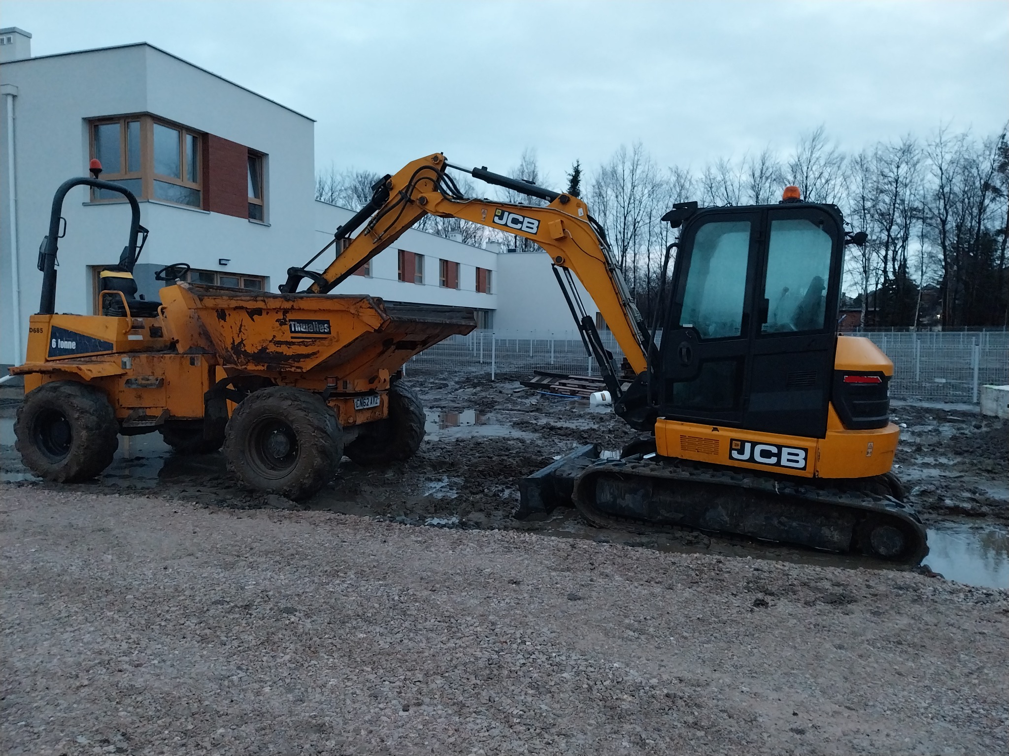Żółta minikoparka JCB i wywrotka Thwaites w błotnistym terenie budowy, na tle nowoczesnego budynku z płaskim dachem i drzew bez liści.
