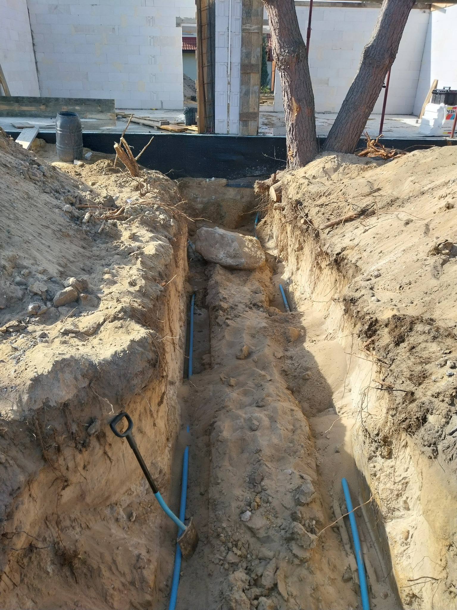 Wykopany rów w piaszczystej ziemi z ułożonymi niebieskimi rurami, widoczna łopata wbita w ścianę wykopu, w tle fundamenty budynku.