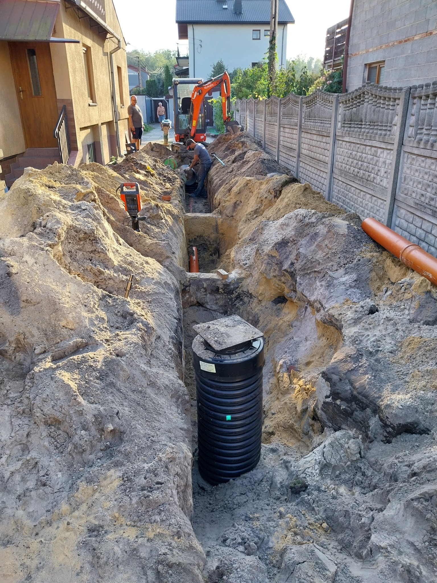 Montaż czarnego szamba z pokrywą w wykopie; w tle pomarańczowa minikoparka i pracujący robotnik, obok wykopu leżąca pomarańczowa rura