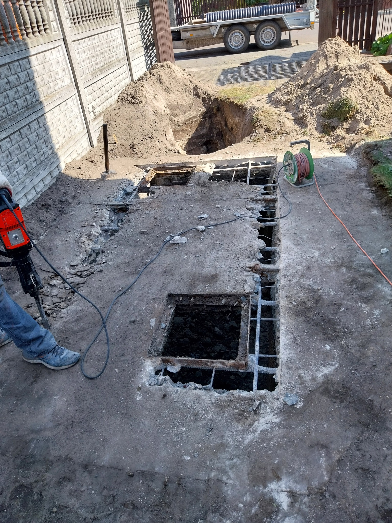 Prace ziemne: Wykopy pod konstrukcję żelbetową z widocznym zbrojeniem, obok leży kabel zasilający i sterta ziemi, w tle ogrodzenie posesji.