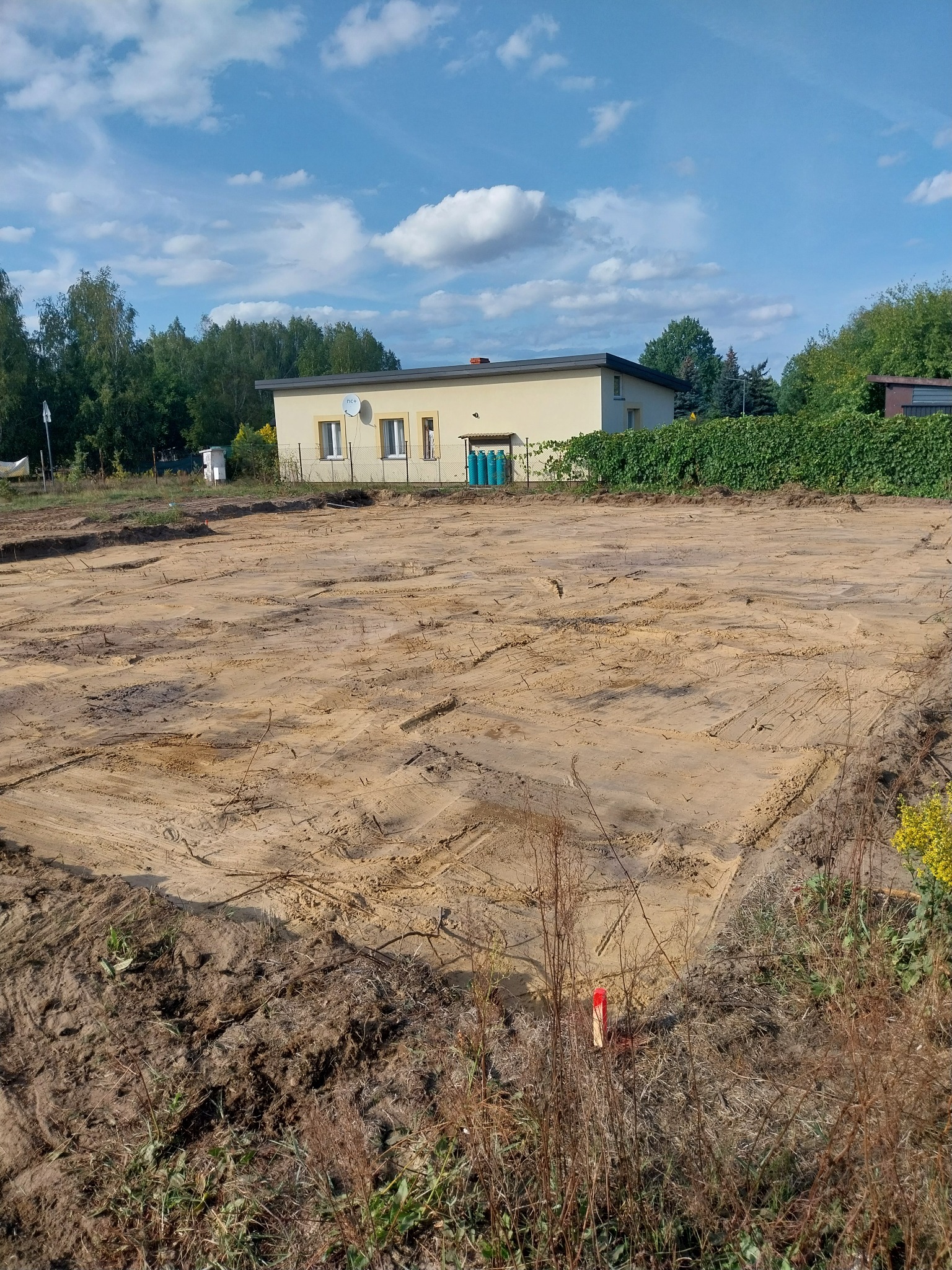 Przygotowany, wyrównany teren pod budowę, widoczne ślady po maszynach, w tle dom jednorodzinny i zieleń.