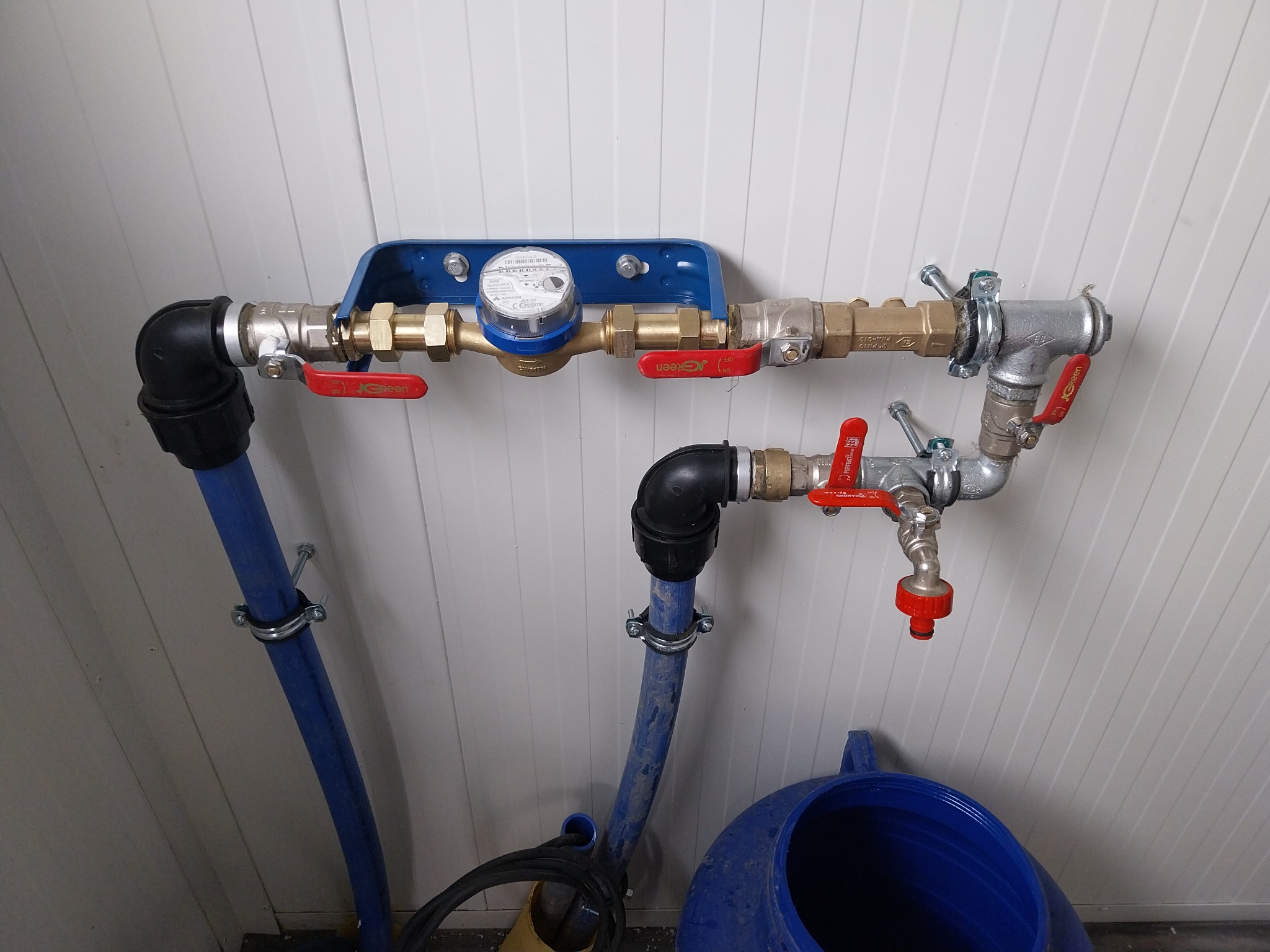 Kompletne przyłącze wodno-kanalizacyjne z licznikiem, zaworami i niebieskimi rurami, zamontowane na białej ścianie z paneli. Widoczne złącza i elementy instalacji.