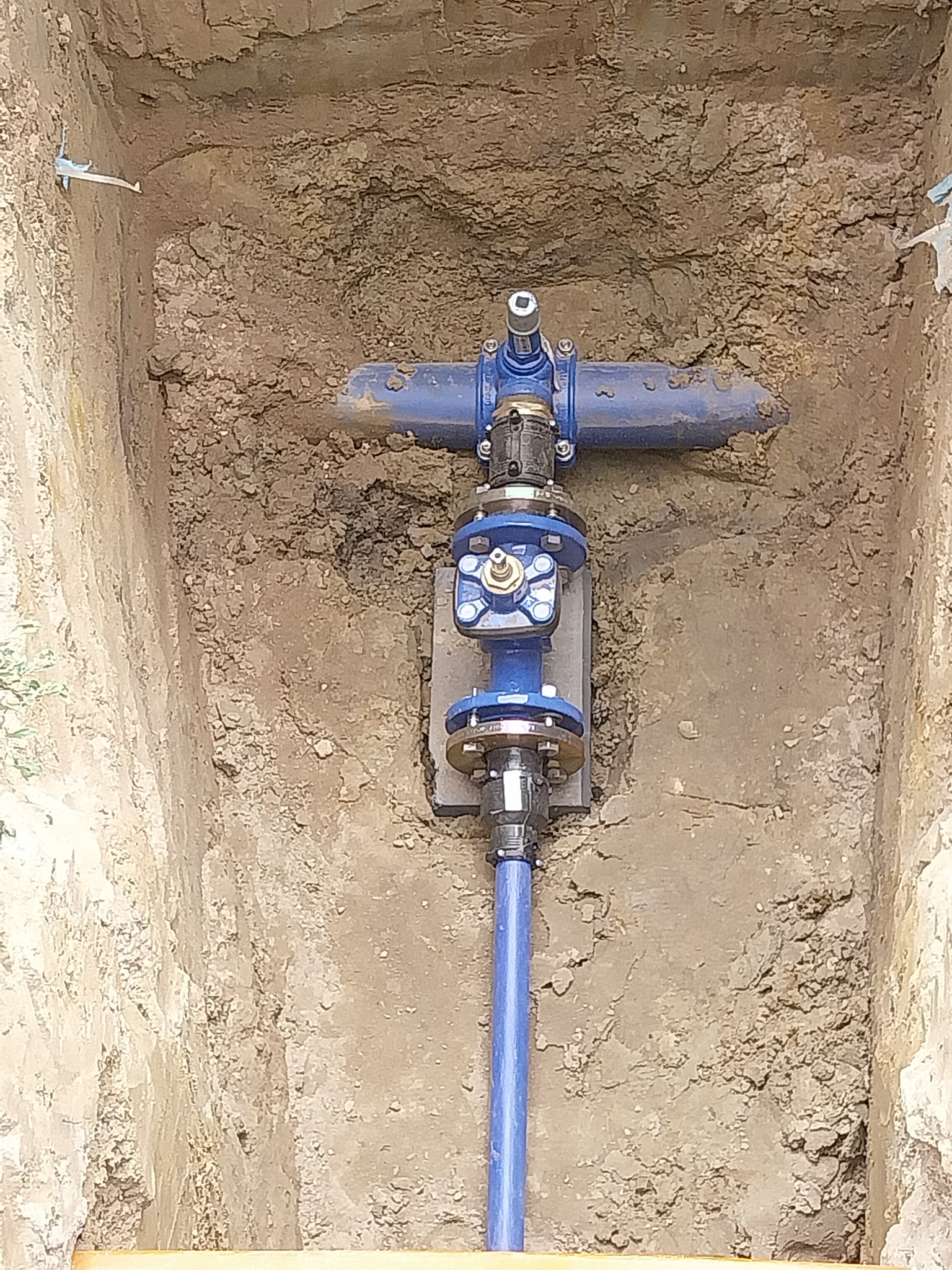 Niebieska instalacja wodociągowa z zaworami i złączkami w wykopie ziemnym, widok z góry. Elementy instalacji są nowe i czyste, kontrastują z surową ziemią.