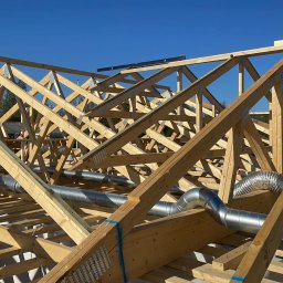 Bro BUD Piotr Kucia - Konstrukcja drewniana dachu w trakcie budowy z widocznymi elementami wentylacji, jasne drewno, metalowe złączki, srebrne rury wentylacyjne, błękitne niebo w tle.