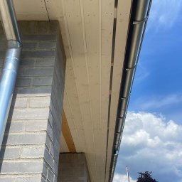 Bro BUD Piotr Kucia - Elewacja budynku w trakcie budowy z widocznym podbitką dachową z paneli PCV, rynną i murem z szarego bloczka betonowego na tle błękitnego nieba.