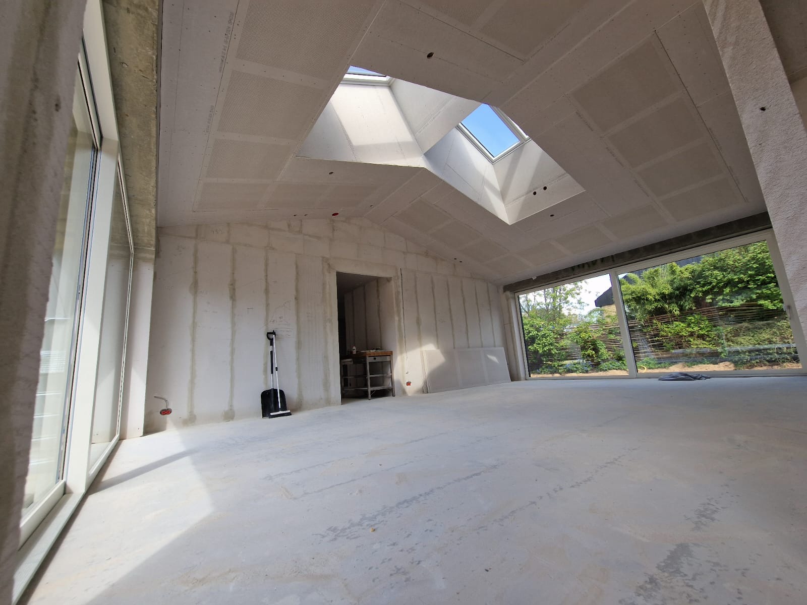 Wnętrze w stanie surowym, ściany i sufit pokryte płytami gipsowo-kartonowymi, widoczne okno dachowe, duże okna z widokiem na zieleń, na podłodze widoczny beton, w rogu stoi czarna zmiotka