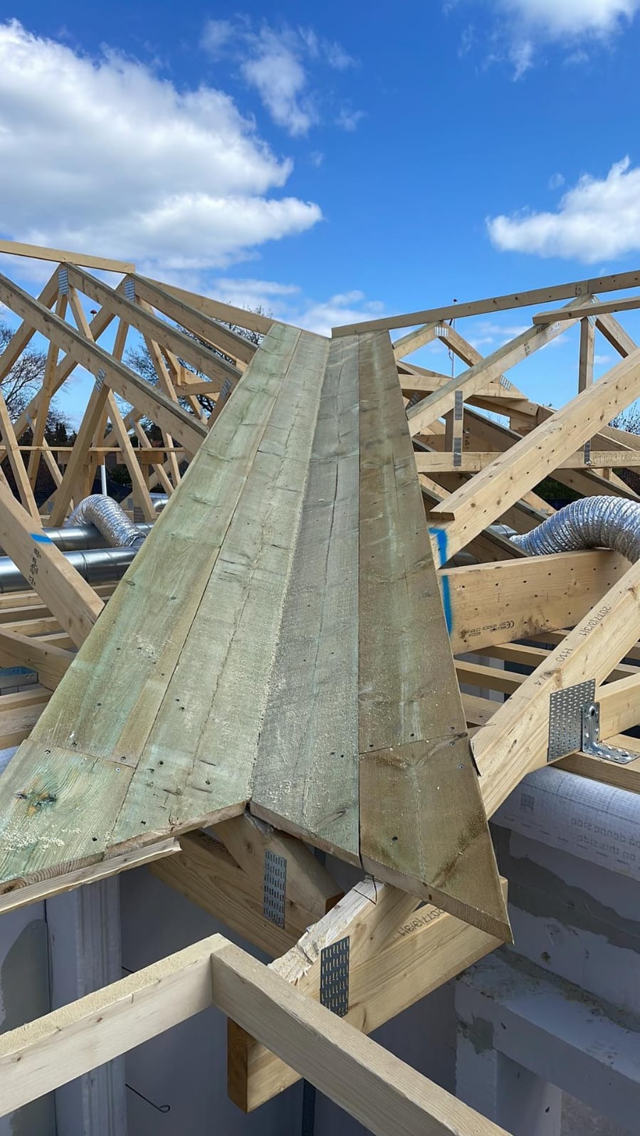 Drewniana konstrukcja dachu w trakcie budowy, widoczne elementy więźby dachowej, połączone metalowymi płytkami, z fragmentem poszycia z desek, na tle błękitnego nieba z chmurami.