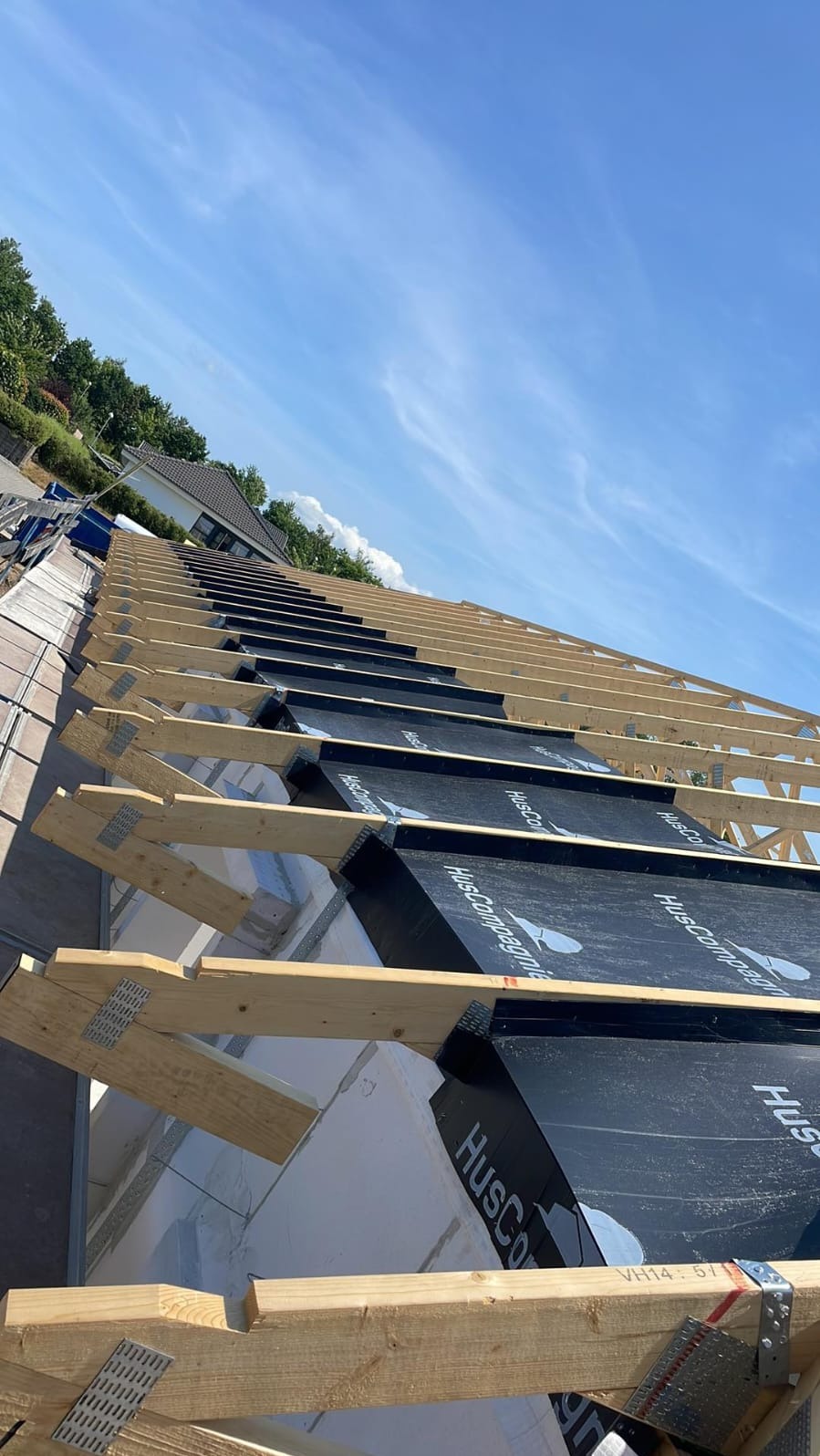 Konstrukcja dachu z widocznymi krokwiami, membraną dachową z nadrukiem HusCompagnie, mocowana metalowymi łącznikami, na tle błękitnego nieba.