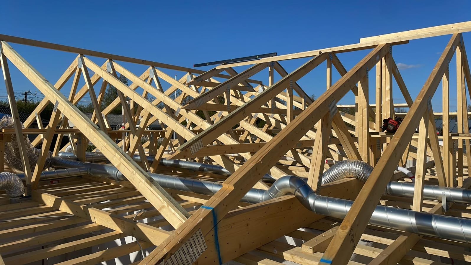Konstrukcja drewniana dachu w trakcie budowy z widocznymi elementami wentylacji, jasne drewno, metalowe złączki, srebrne rury wentylacyjne, błękitne niebo w tle.