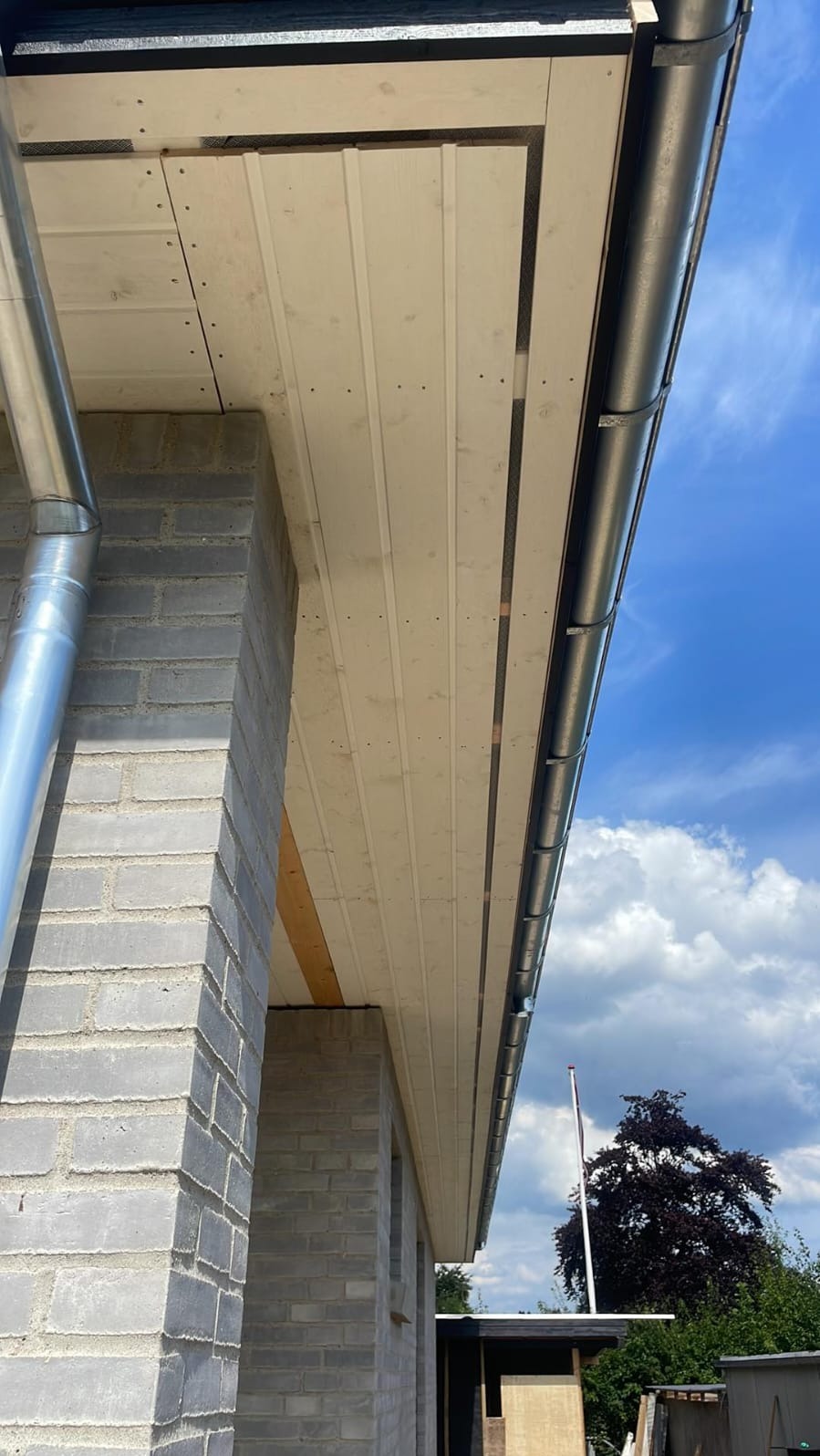 Elewacja budynku w trakcie budowy z widocznym podbitką dachową z paneli PCV, rynną i murem z szarego bloczka betonowego na tle błękitnego nieba.