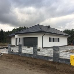 Olomk Waldemar Schulz - Budowa ogrodzenia z szarych bloczków betonowych przed nowoczesnym domem z szarą bramą garażową i kostką brukową na podjeździe, widoczna skrzynka gazowa.