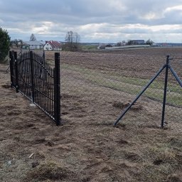 Olomk Waldemar Schulz - Montaż czarnej, metalowej bramki furtki w siatce ogrodzeniowej na tle zaoranego pola i zabudowań wiejskich pod pochmurnym niebem.