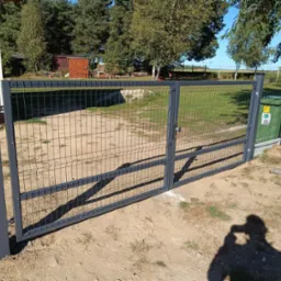 Szara, metalowa brama panelowa, dwuskrzydłowa, z widocznym słupkiem i narzędziami montażowymi obok. Tło: trawa, drzewa, budynek. Cień osoby robiącej zdjęcie.