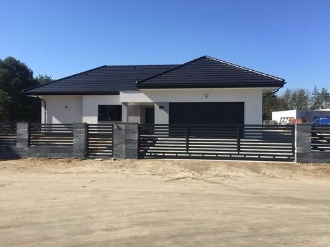 Nowoczesny dom jednorodzinny z czarną, poziomą, metalową konstrukcją ogrodzeniową z szarymi, kamiennymi słupkami, widok z przodu w słoneczny dzień.