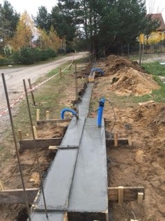 Wąski fundament betonowy w drewnianym szalunku, przygotowany pod budowę ogrodzenia, widoczne rury instalacyjne wystające z ziemi.