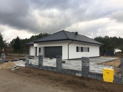 Budowa ogrodzenia z szarych bloczków betonowych przed nowoczesnym domem z szarą bramą garażową i kostką brukową na podjeździe, widoczna skrzynka gazowa.
