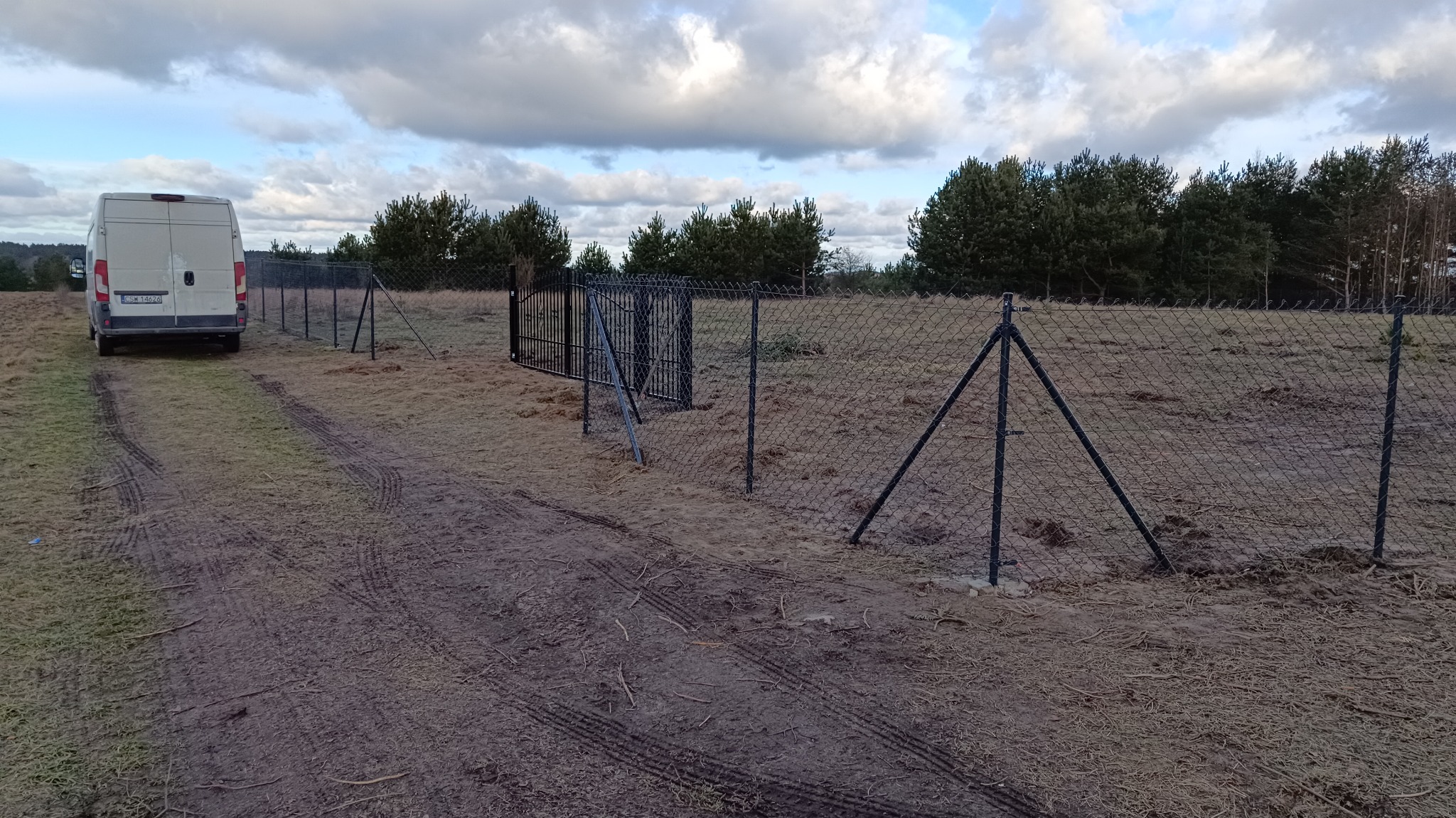 Montaż siatki ogrodzeniowej z bramą wjazdową na działce, widoczny biały samochód dostawczy i ślady opon na ziemi.