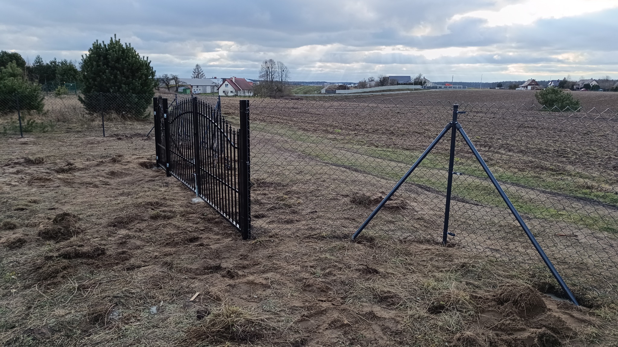 Montaż czarnej, metalowej bramki furtki w siatce ogrodzeniowej na tle zaoranego pola i zabudowań wiejskich pod pochmurnym niebem.