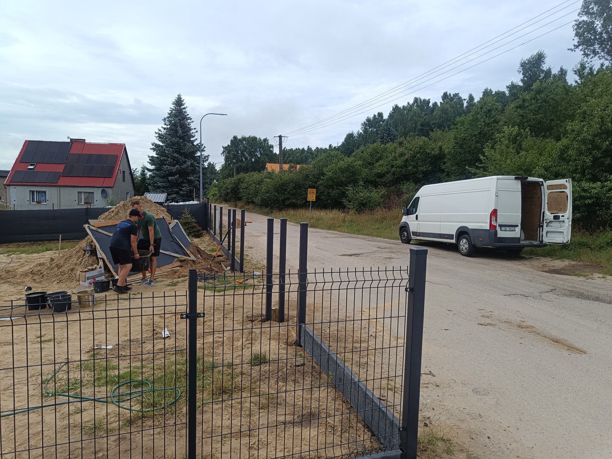 Montaż ogrodzenia panelowego z podmurówką: Dwóch mężczyzn pracuje przy instalacji, widoczny van z otwartymi drzwiami, dom z panelami słonecznymi w tle.