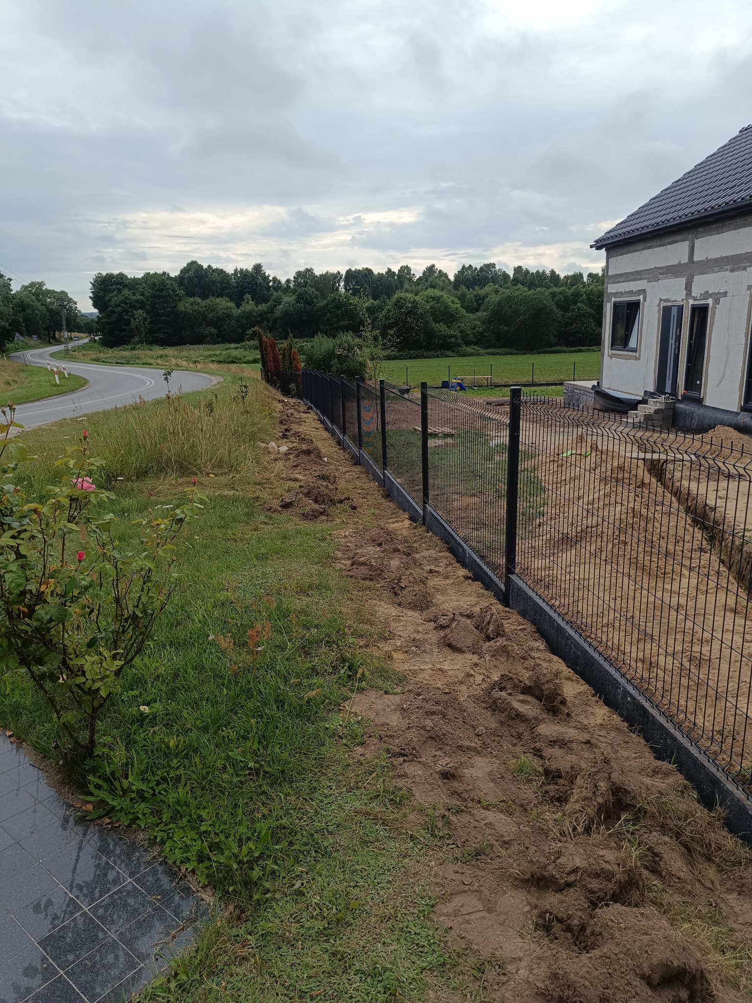 Czarny panel ogrodzeniowy z podmurówką na nierównym terenie, w tle dom w trakcie budowy i zieleń. Widok z boku na nowo postawione ogrodzenie.