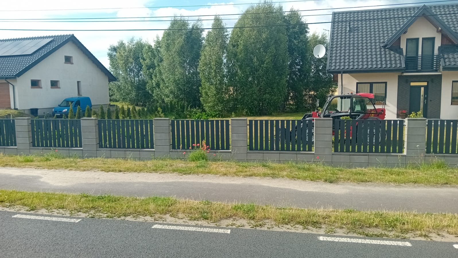 Nowoczesne ogrodzenie z szarych, betonowych słupków i czarnych, pionowych paneli na tle dwóch domów i zieleni, widziane z perspektywy ulicy.