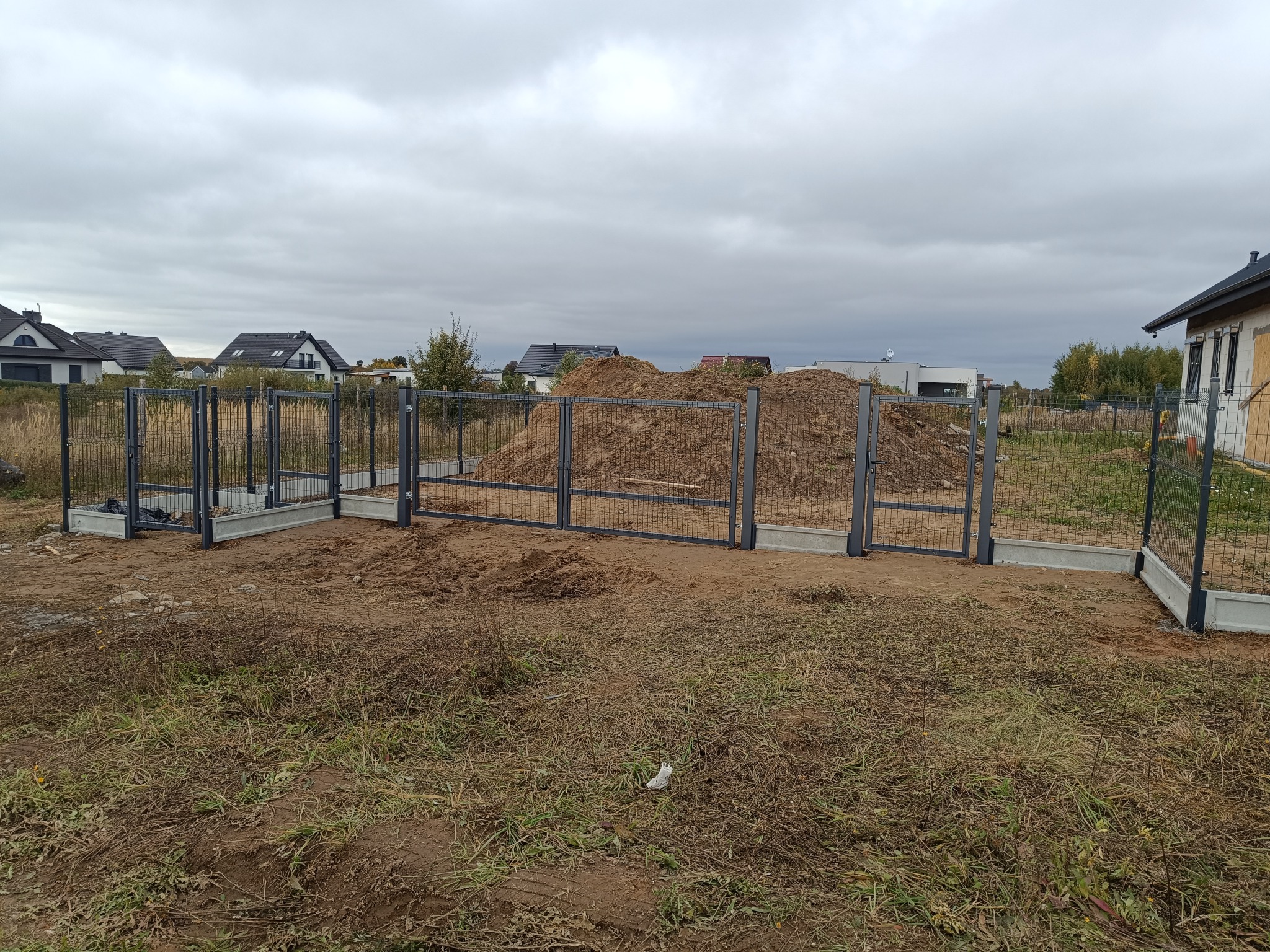 Fragment montażu grafitowego ogrodzenia panelowego na betonowej podmurówce, z ziemią w tle i widoczną zabudową jednorodzinną w oddali.