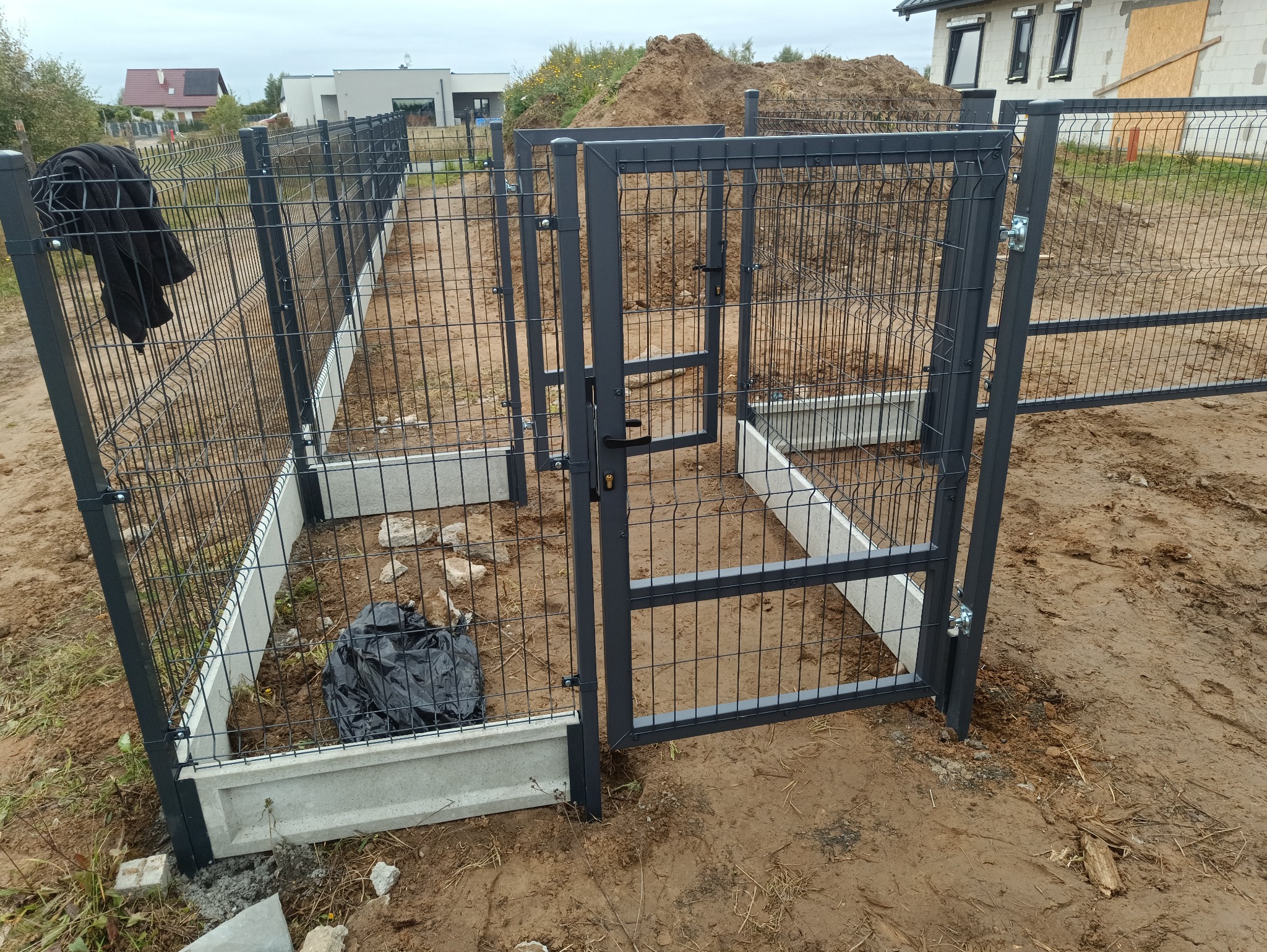 Szara furtka zamykająca ogrodzenie panelowe z podmurówką betonową na działce w trakcie budowy, widok z bliska na zamek i ziemię.